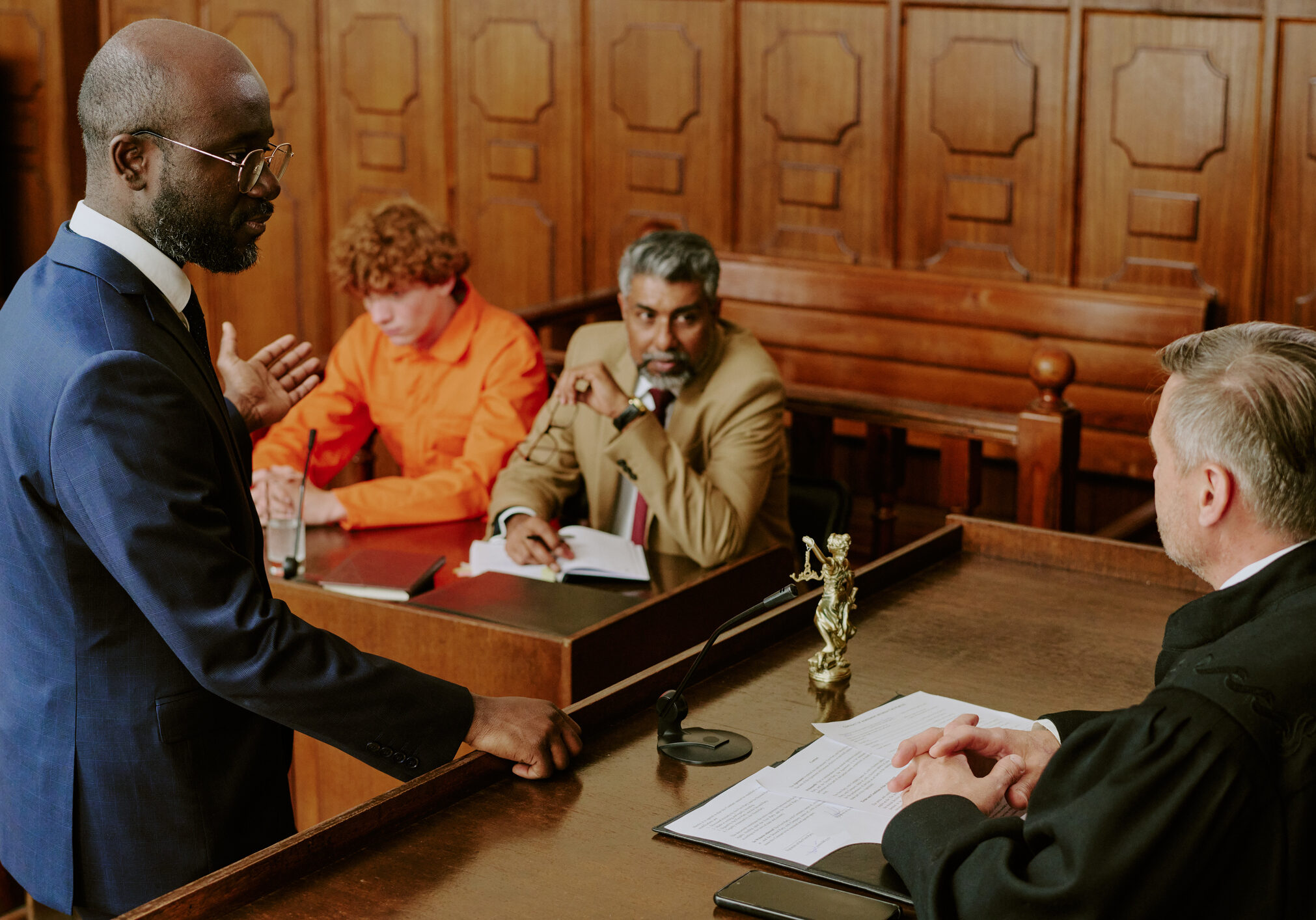 Black man in suit addressing Caucasian male judge in courtroom, while middle aged biracial attorney and young adult Caucasian defendant in orange jumpsuit sitting at table listening